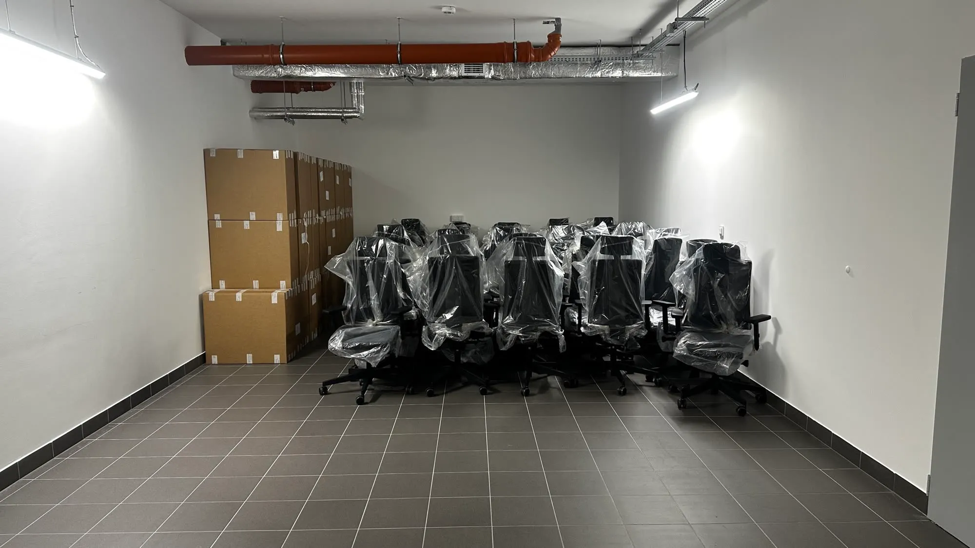 Kilkanaście foteli biurowych owiniętych folią, ustawionych pod ścianą pustego pomieszczenia